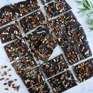 pecan rosemary chocolate bark cut into squares and arranged on marble table with rosemary sprigs in background