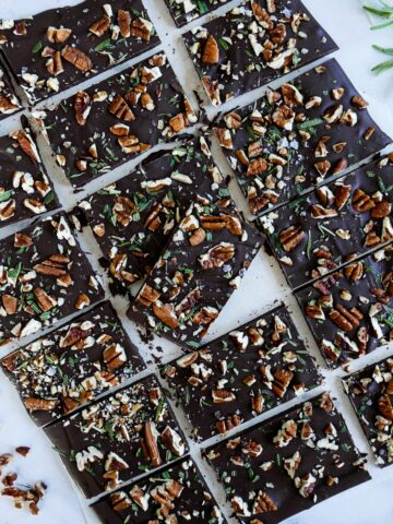 pecan rosemary chocolate bark cut into squares and arranged on marble table with rosemary sprigs in background
