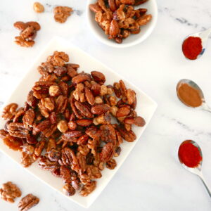 spiced candied nuts arranged on a white plate with nuts scattered around and three spoons full of spices nearby