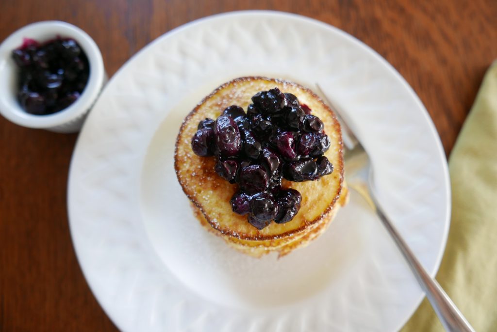 ricotta pancakes with blueberry sauce piled on a white plate with additional blueberry sauce on the side
