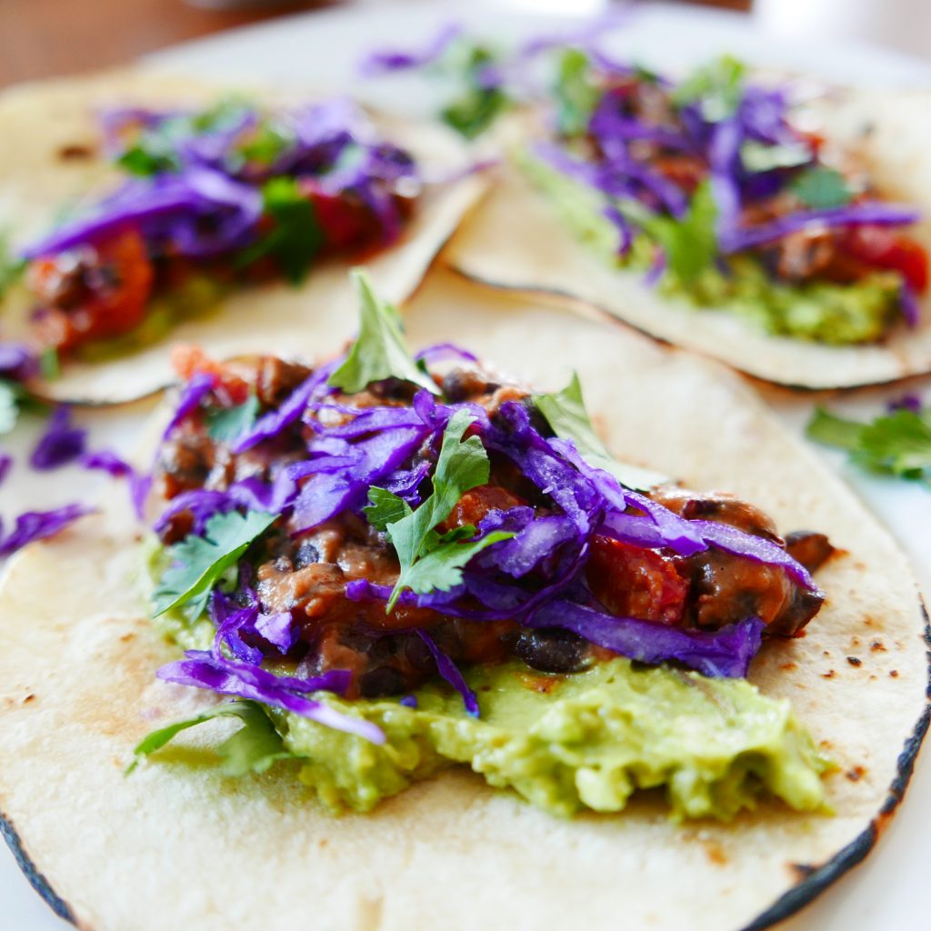 close up of three black bean and cheese tacos garnished with smashed avocado, shredded red cabbage and green cilantro.