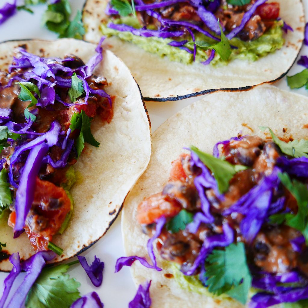 close up of three black bean and cheese tacos garnished with smashed avocado, shredded red cabbage and green cilantro.