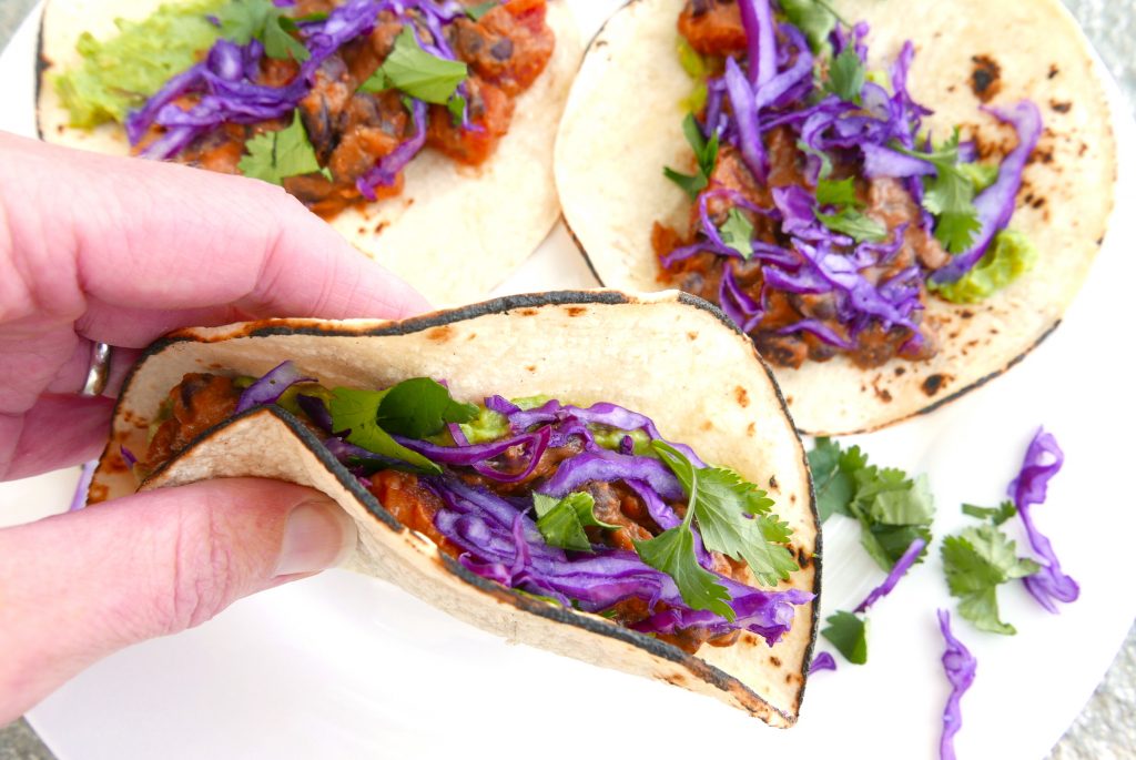 white hand holding folded black bean and cheese taco with red cabbage and green cilantro. Two additional tacos are in the background sitting on a white plate.