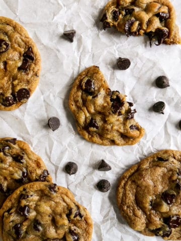 five brown butter chocolate chip cookies arranged on parchment paper with chocolate chips.