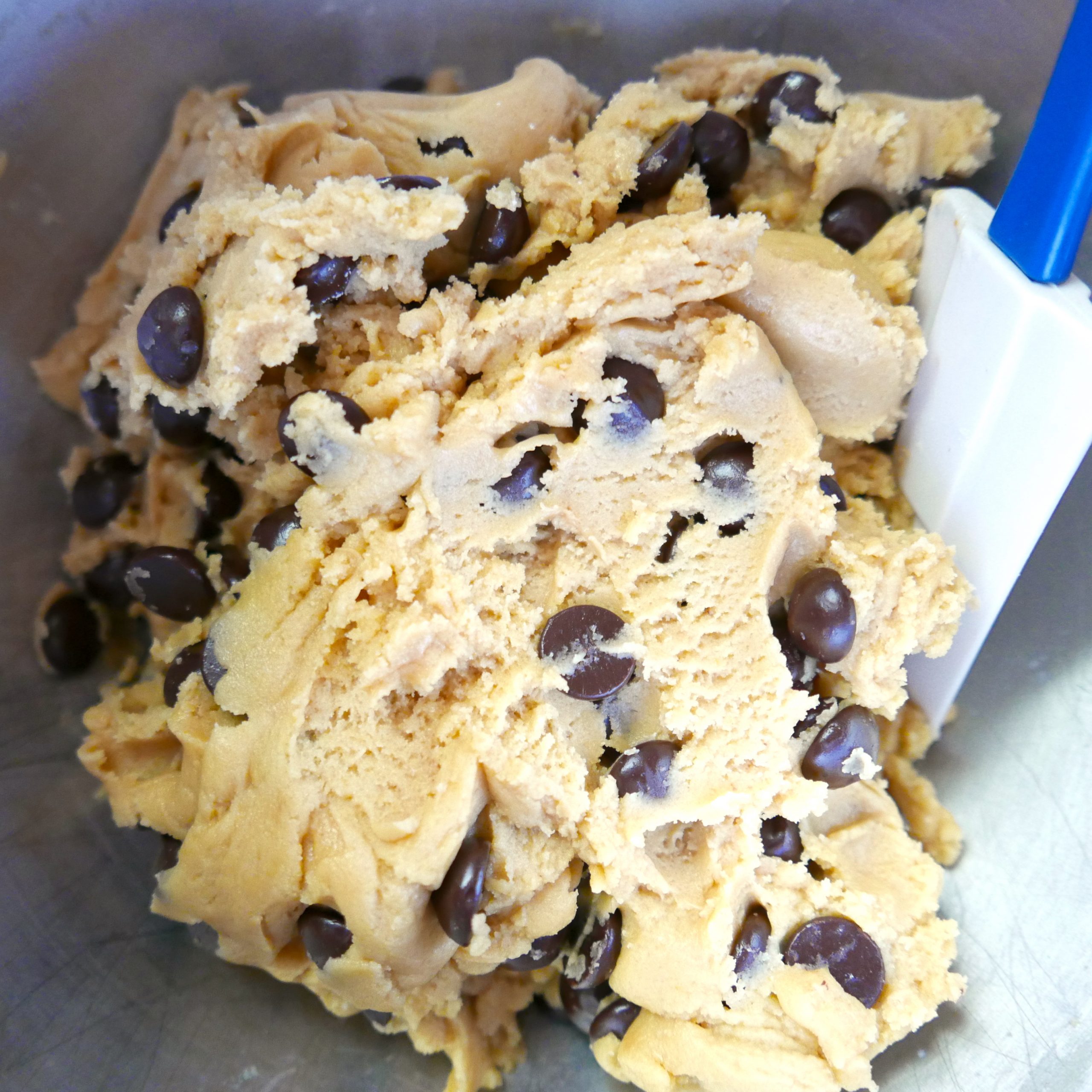 close up of chocolate chip cookie dough in a silver bowl with white rubber spatula.