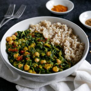 bowl of chickpea spinach curry with brown rice resting on a white napkin with forks and spices in the background