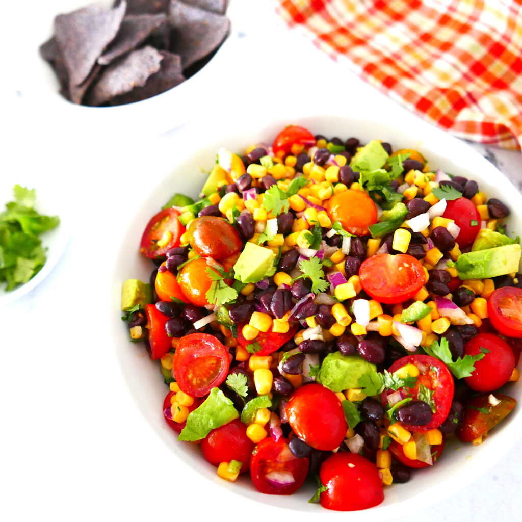 white bowl containing corn avocado black bean salsa with small bowl of tortilla chips in background and small bowl of cilantro next to it