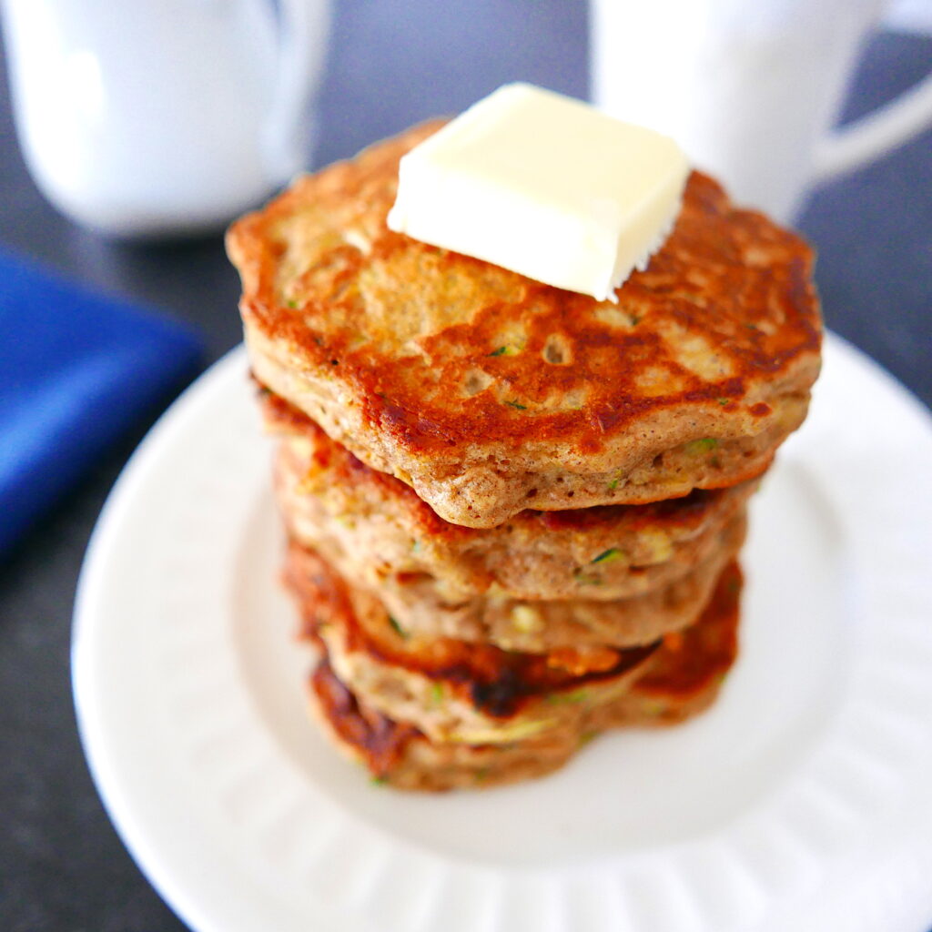 a large stack of zucchini pancakes with a square of butter on top arranged on a white plate with white coffee cup in background and blue napkin next to it