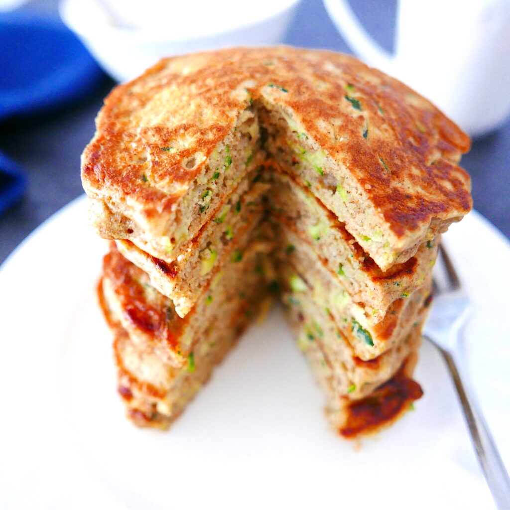 a large stack of zucchini bread pancakes with a large piece cut out in front arranged on a white plate with fork