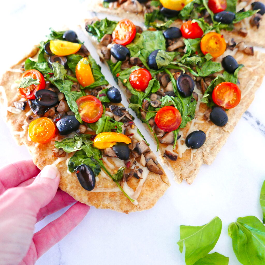 white hand picking up a piece of vegan flatbread pizza with lots of vegetables on top