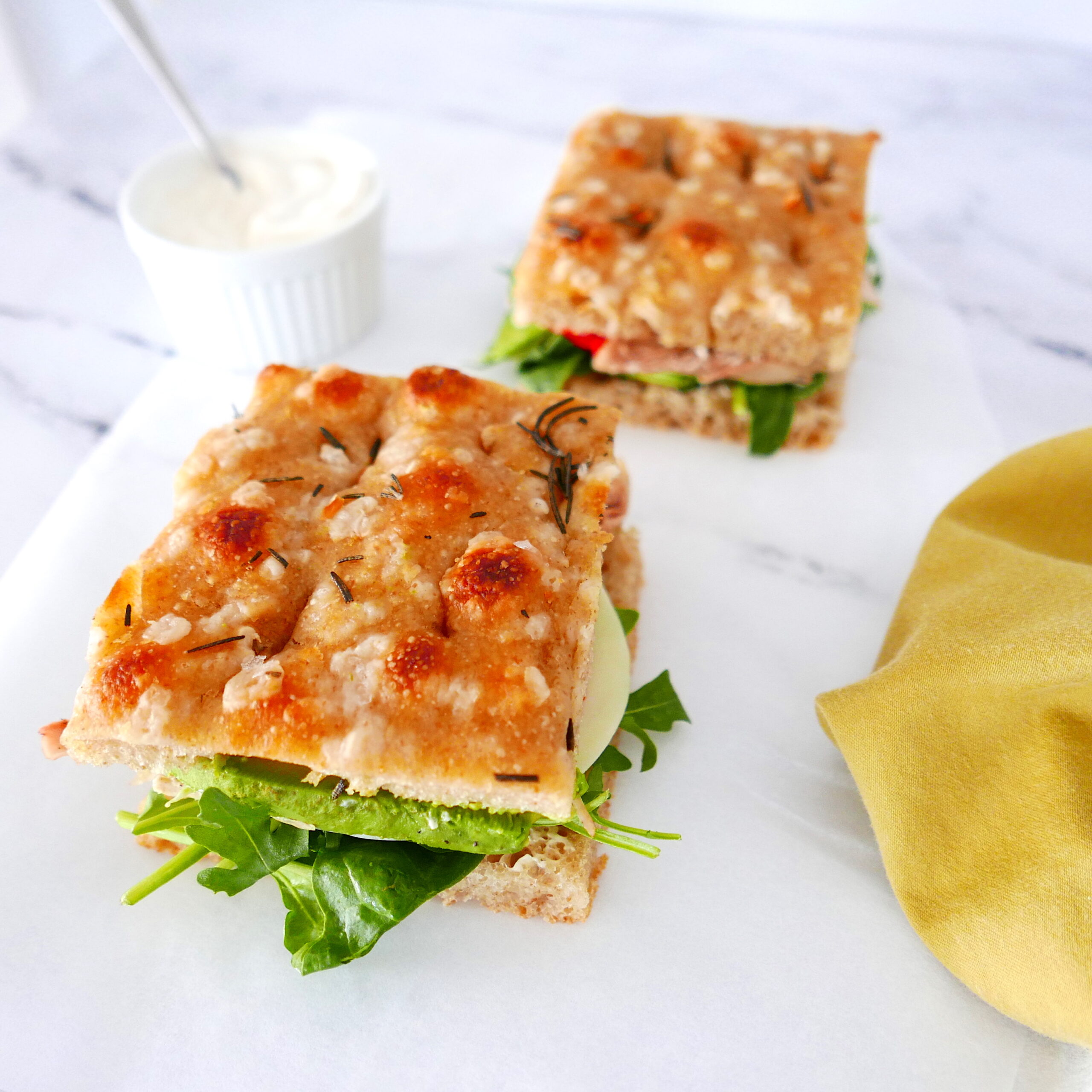 Two focaccia sandwiches on a marble counter with a cup of mayo in the background.