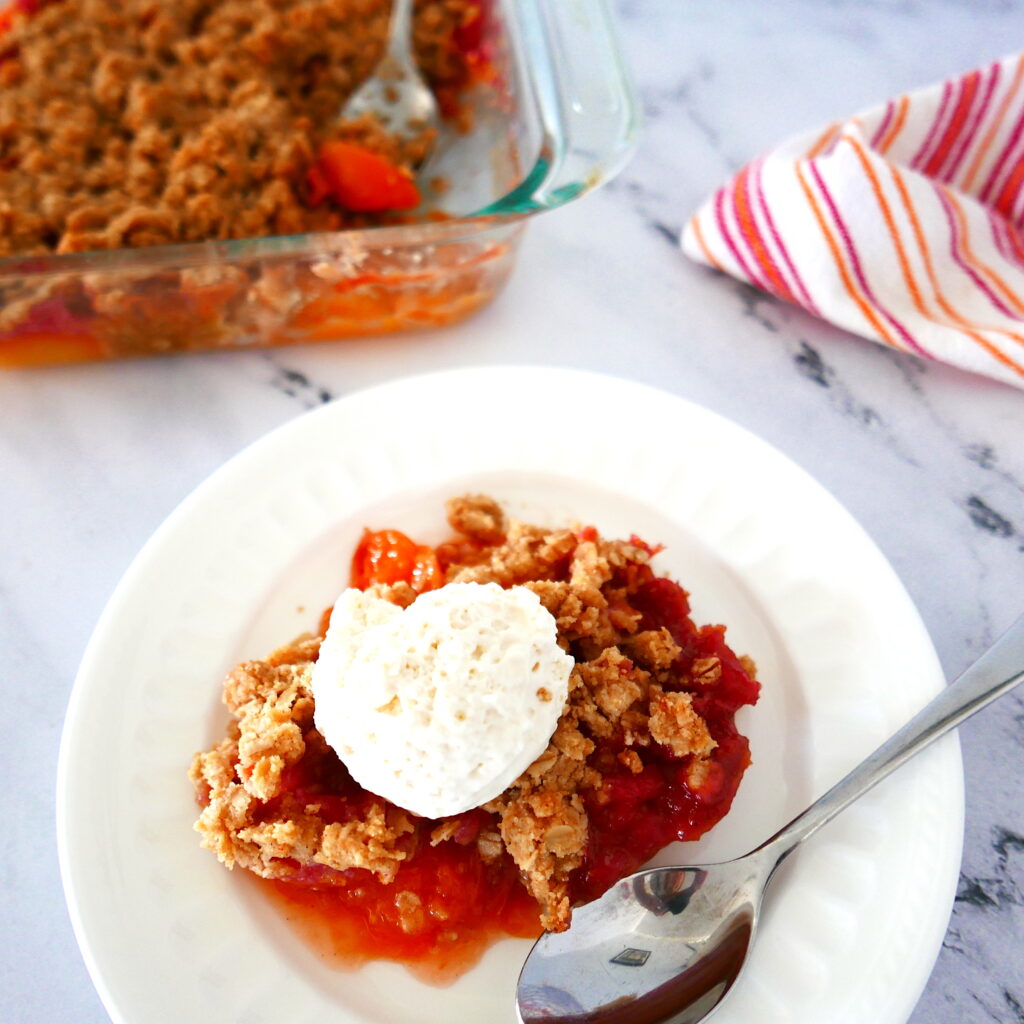 peach crisp with whipped cream and spoon on a white plate with a pan of peach crisp in the background