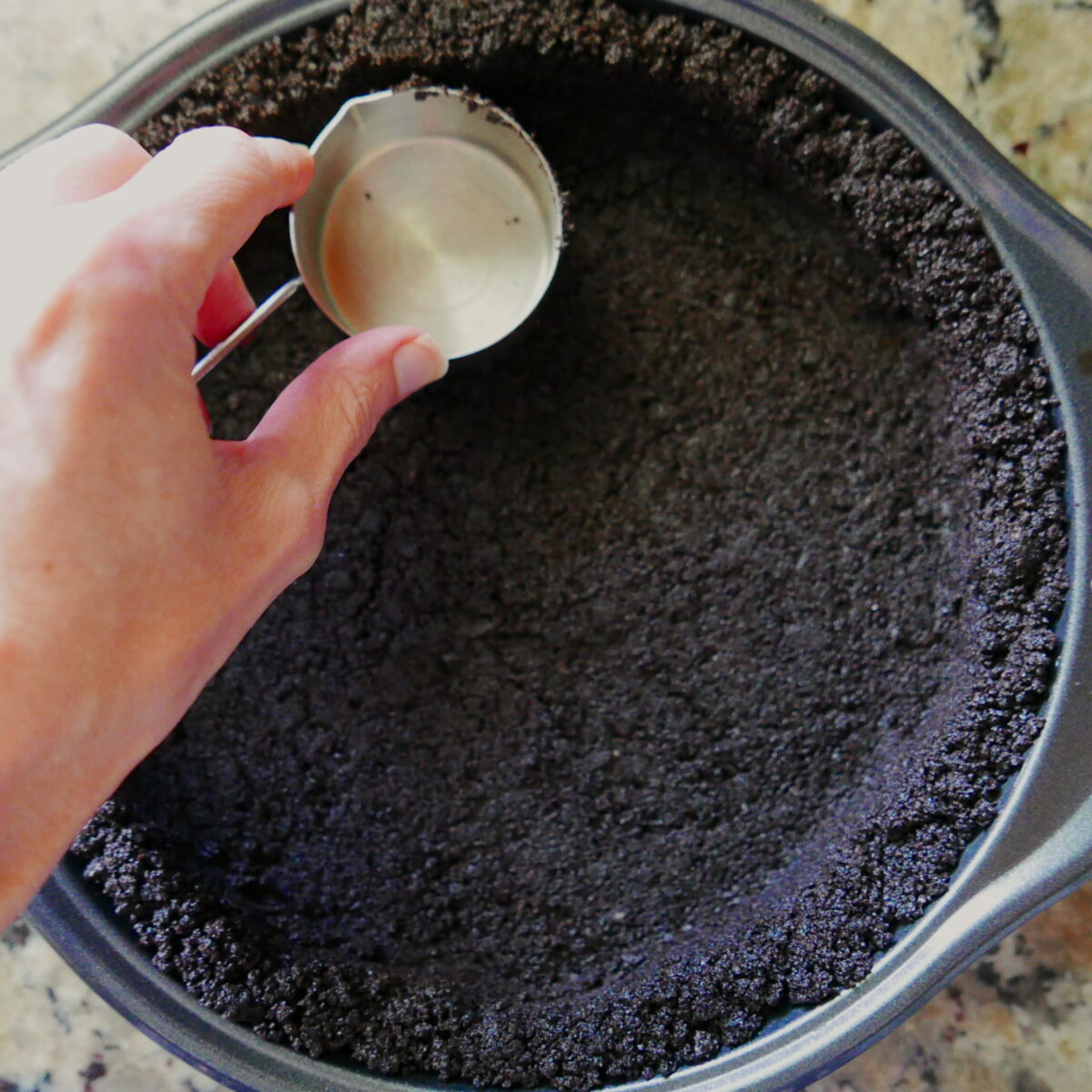 White hand pressing measuring cup into chocolate cookie crust in a pie pan.