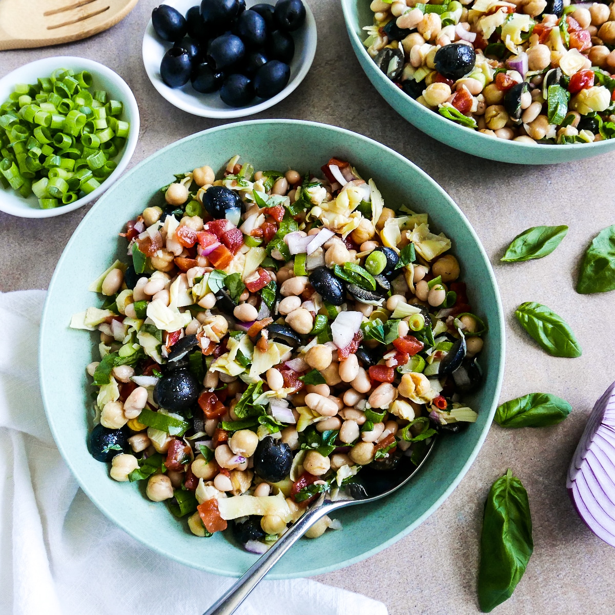 Bowl of tuscan bean salad with a spoon, fresh basil, and a white napkin.