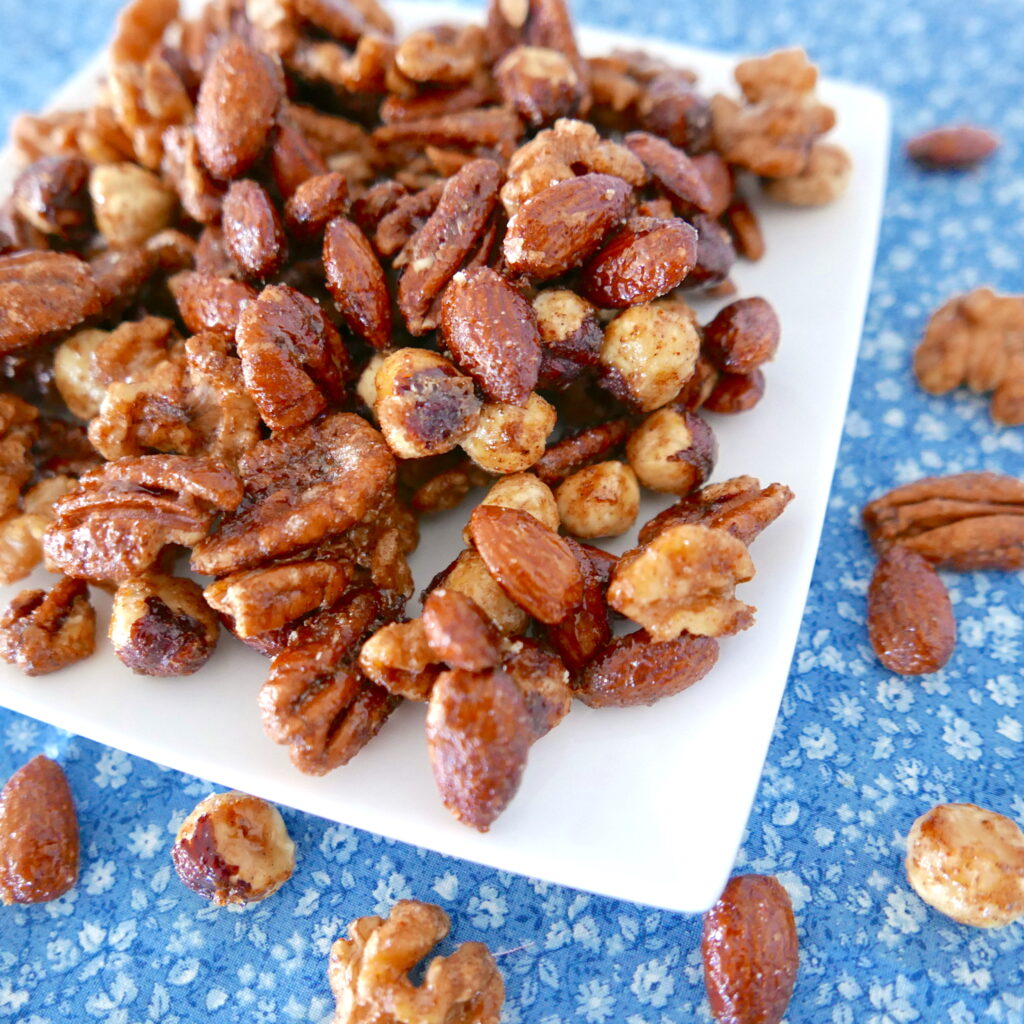 spiced candied mixed nuts arranged on a white plate with nuts scattered around on a blue tablecloth
