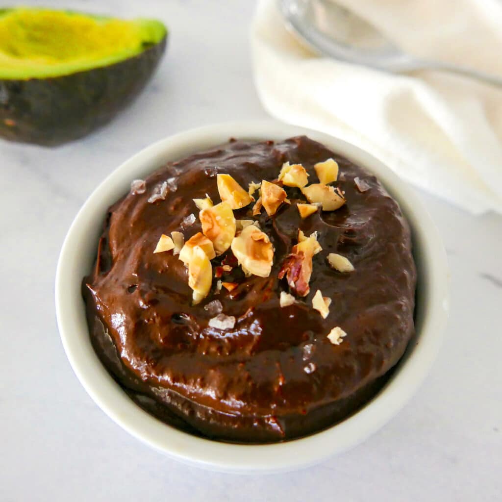 chocolate avocado pudding with chopped hazelnuts in a white cup with spoon in background