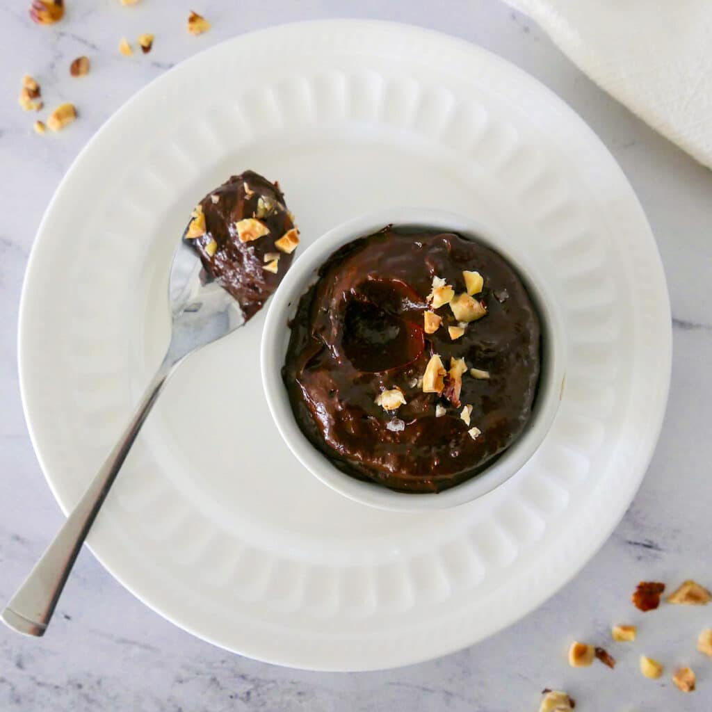 chocolate avocado pudding no bake in a small white cup with spoon and chopped hazelnuts