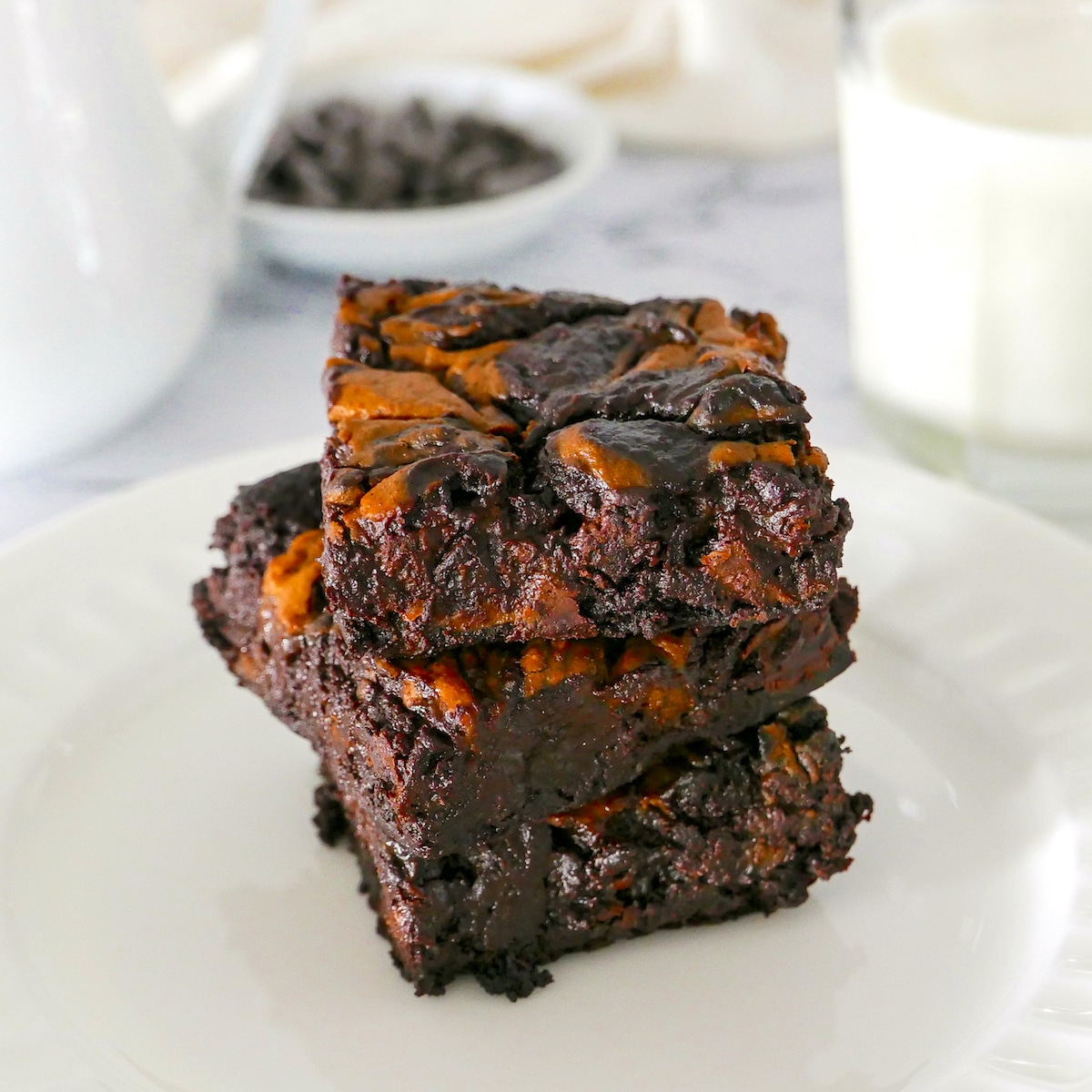three vegan brownies stacked on a white plate with glass of milk in background