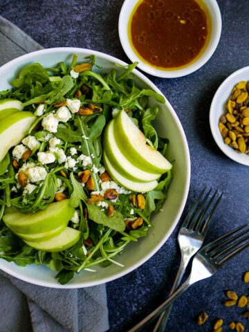 Apple salad in white bowl with two forks and pistachios in a cup