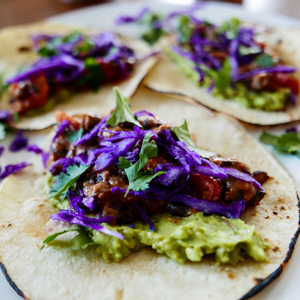 Three tacos on a white plate garnished with cilantro, avocado, and red cabbage.
