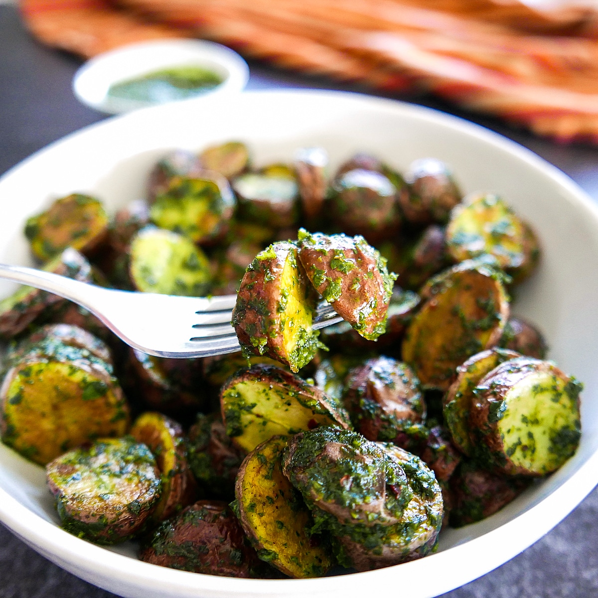 Fork lifting up bite of red potatoes with chimichurri sauce from a white bowl.