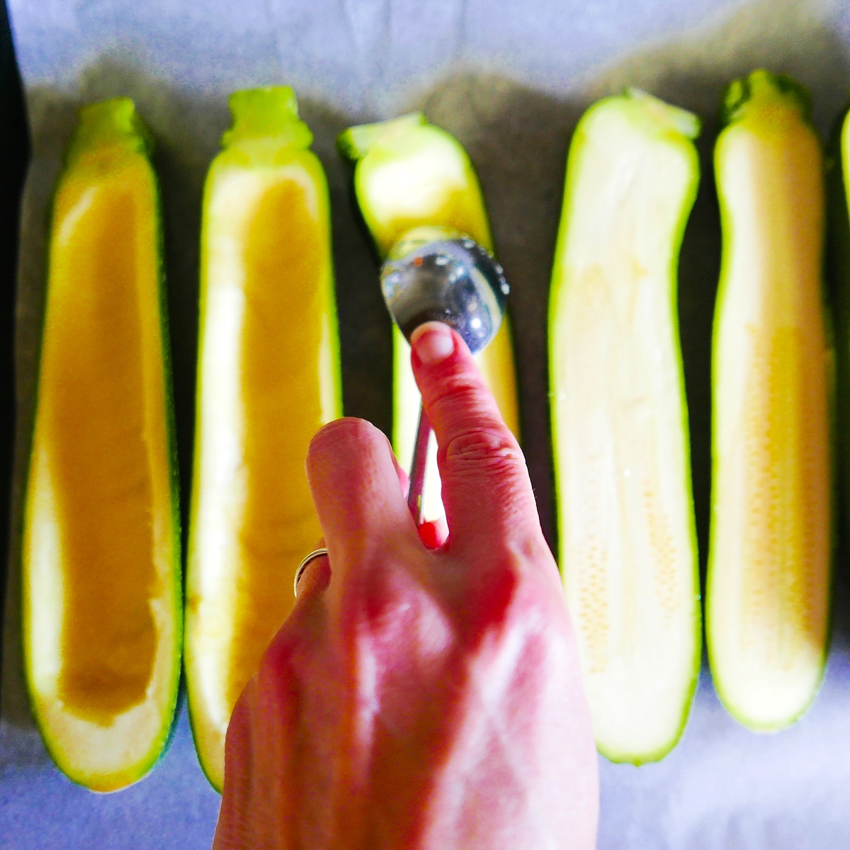 white hand using a spoon to scoop out zucchini on a baking sheet