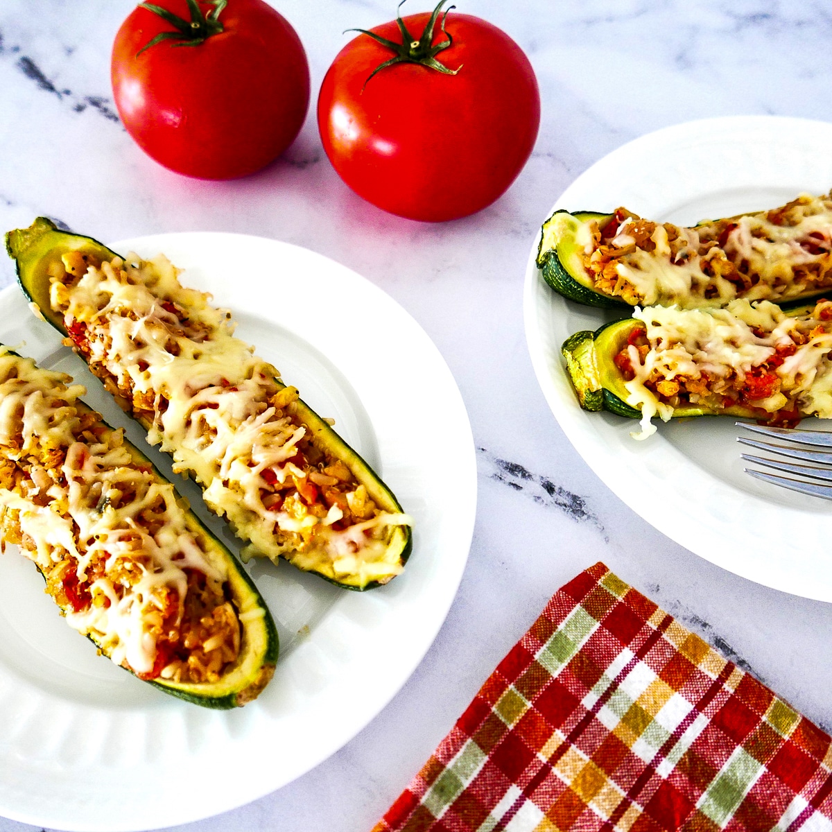 four stuffed zucchini on two white plates with tomatoes in the background