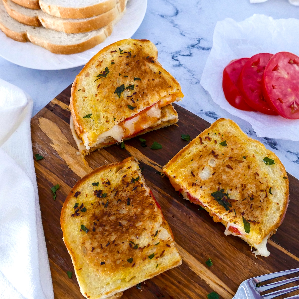 Three pieces of grilled cheese on cutting board with tomato and sourdough slices in background.