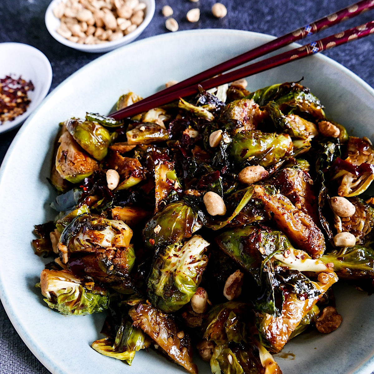 kung-pao-brussels-sprouts-with-chopsticks.jpg kung pao brussels sprouts garnished with peanuts and chopsticks in a white bowl with cup of peanuts in background