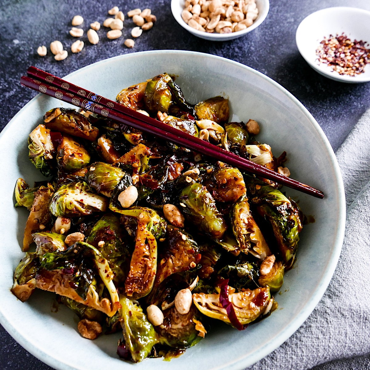 kung-pao-brussels-sprouts-with-peanuts.jpg kung pao brussels sprouts in a white bowl with chopsticks and cup of red pepper flakes in background