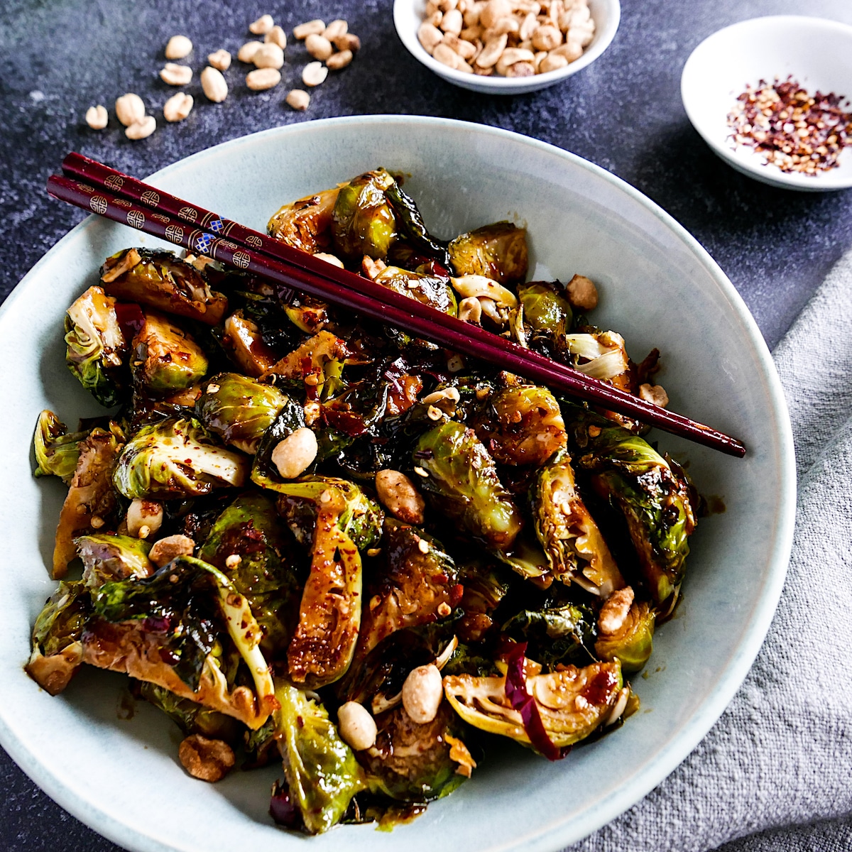 Kung pao Brussels sprouts in a white bowl with chopsticks and cup of red pepper flakes.