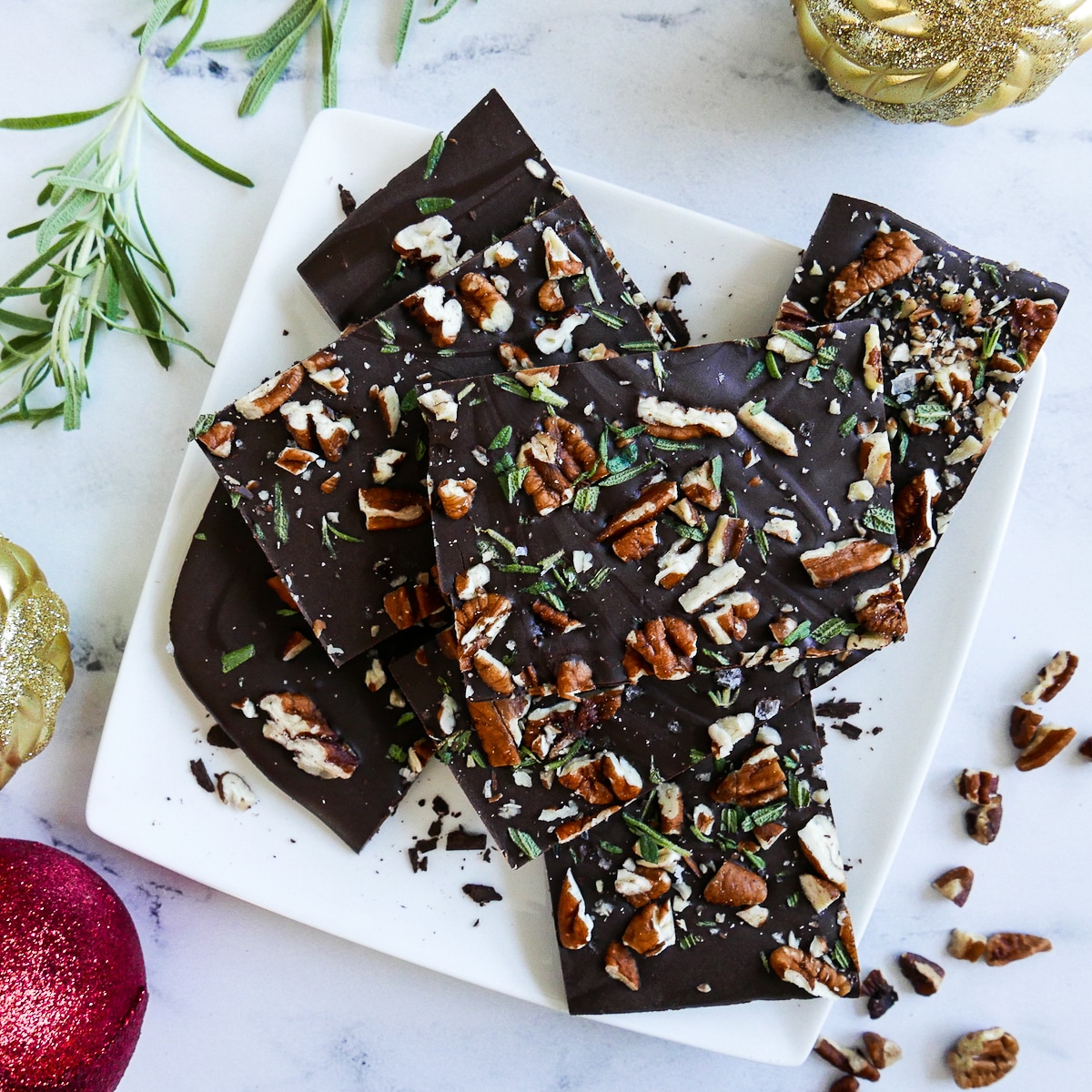 chocolate-pecan-rosemary-bark-on-plate.jpg chocolate pecan rosemary bark cut into squares and arranged on a white plate with chopped pecans and rosemary next to plate
