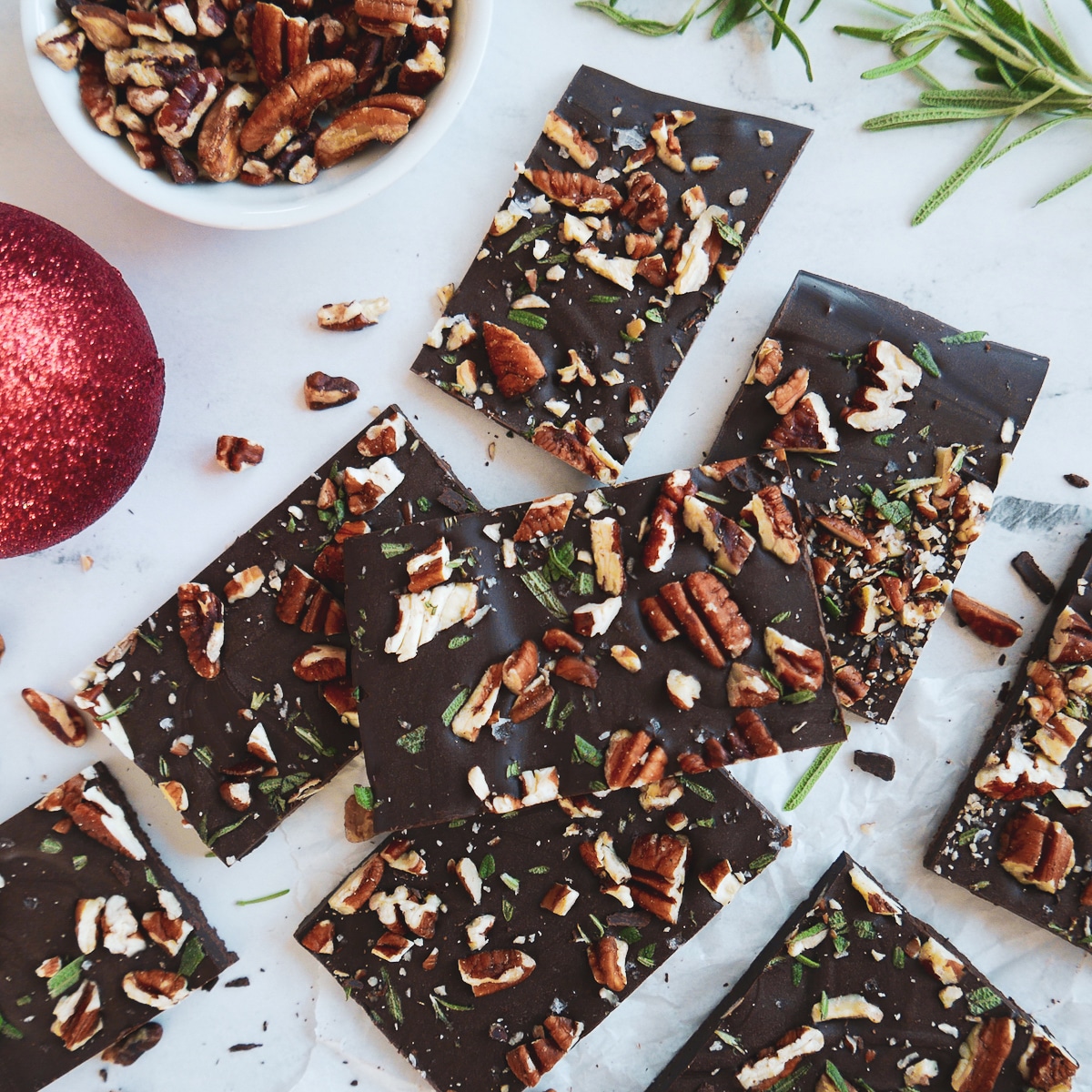 chocolate-pecan-bark.jpg chocolate pecan rosemary bark arranged on a white table with rosemary springs and chopped pecans around