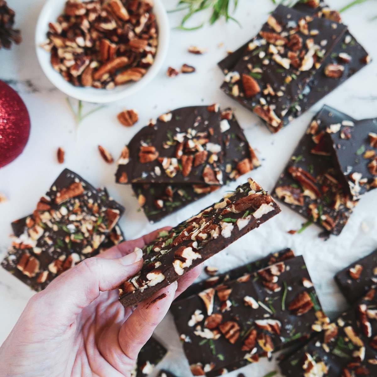 chocolate-pecan-bark-in-hand.jpg chocolate pecan rosemary bark arranged on a marble table with hand holding up one piece and cup of chopped pecans in background