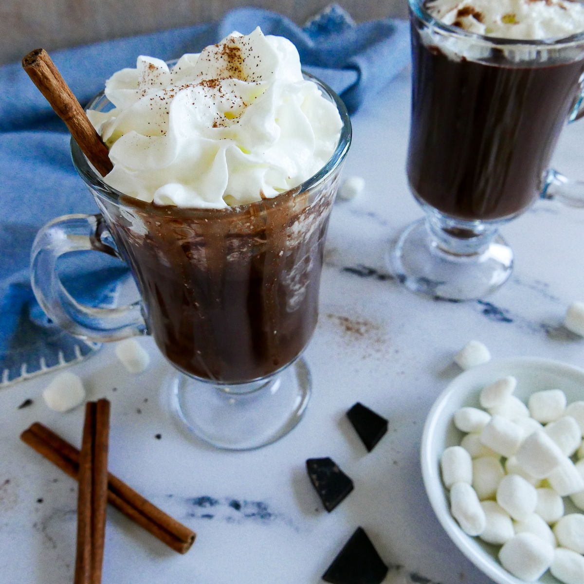 two mugs of oat milk hot chocolate garnished with whipped cream and a cinnamon stick.
