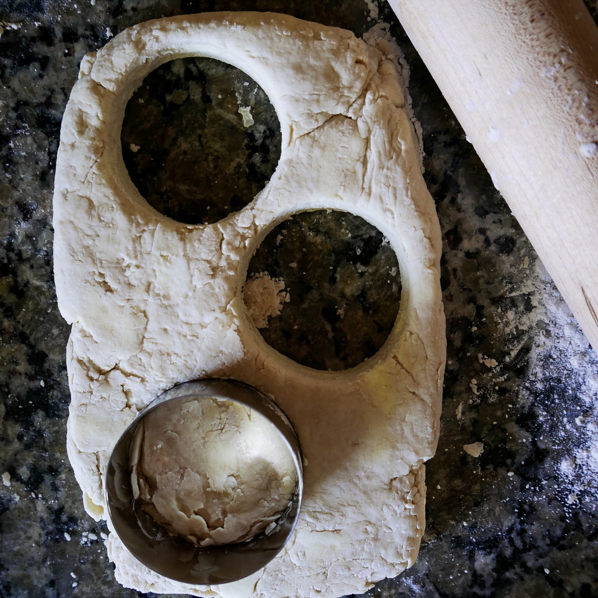 Biscuit cutter cutting out three biscuits with rolling pin next to them.