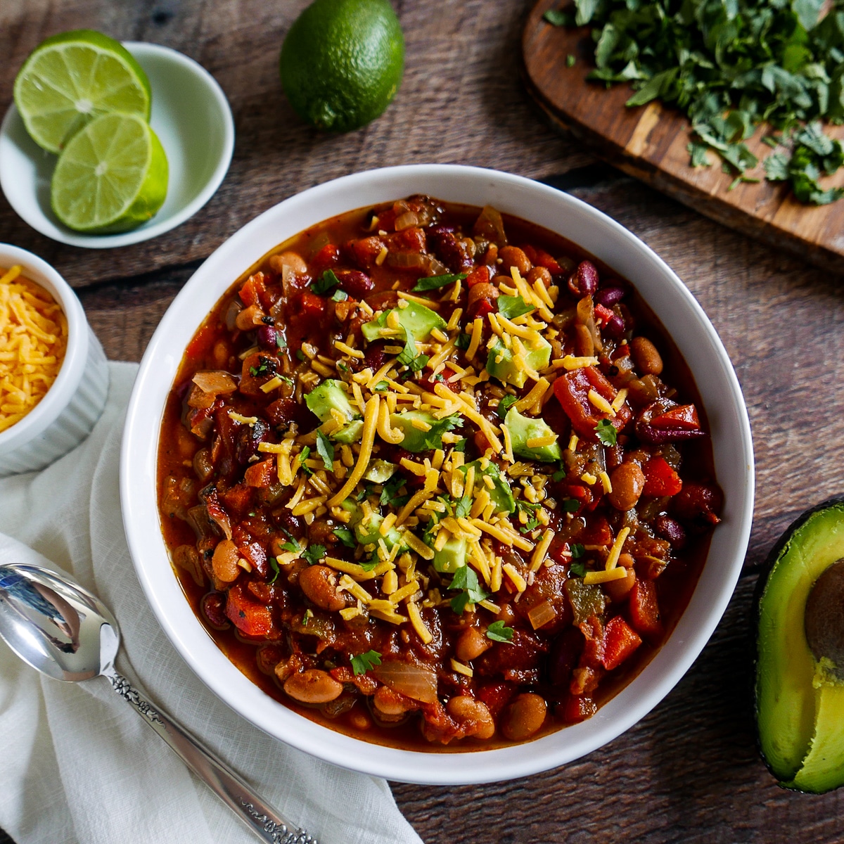 bowl of vegetarian chili with spoon, avocado, cheese, cilantro, and lime.