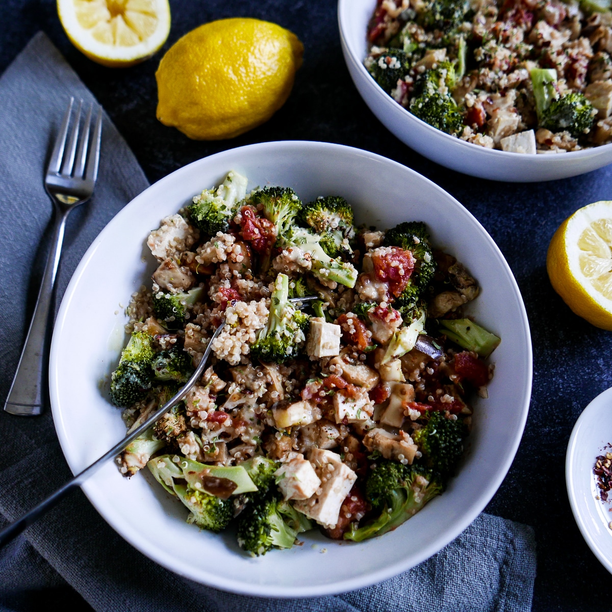 tahini quinoa bowl with eggplant, broccoli, and tofu with two forks and lemons.