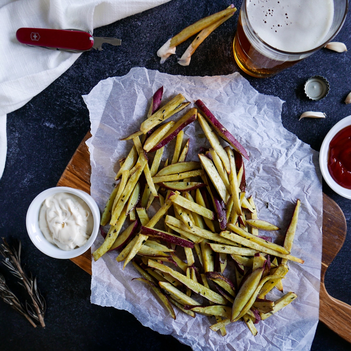 Sweet potato fries on a wooden cutting board with beer and condiments nearby.