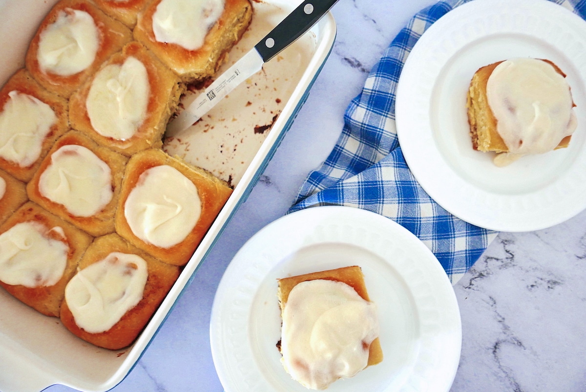 gooey-cinnamon-rolls.jpg two gooey cinnamon rolls on white plates with pan of rolls next to them with a knife.