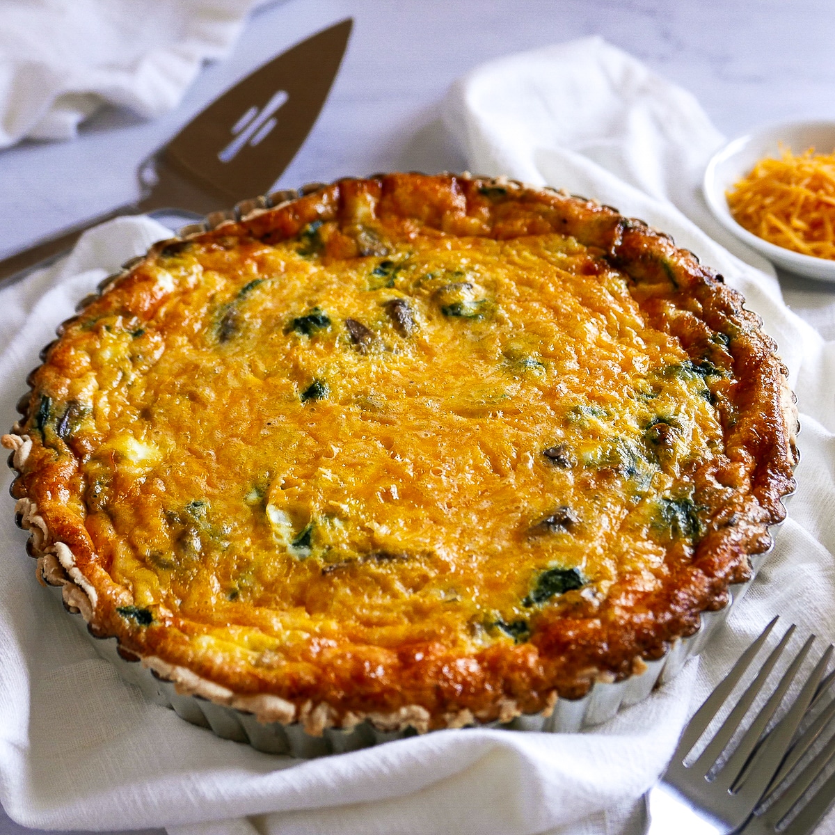 Baked spinach quiche in a tart pan with pie server, napkin, and cheese in background.