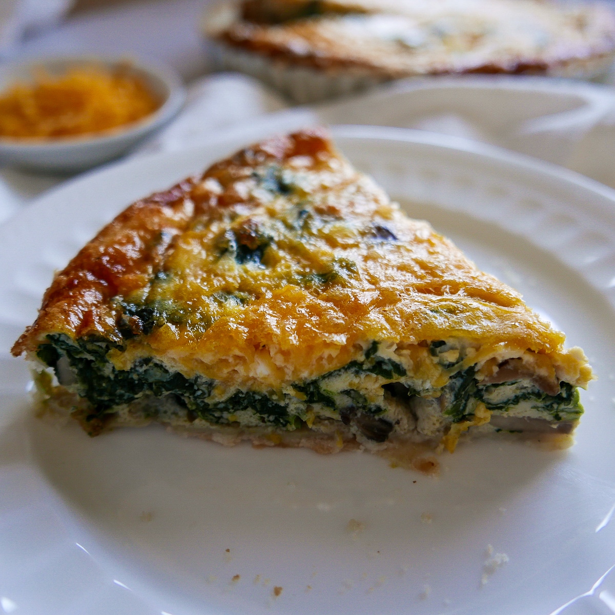 One piece of quiche on a white plate with pan of quiche in background.