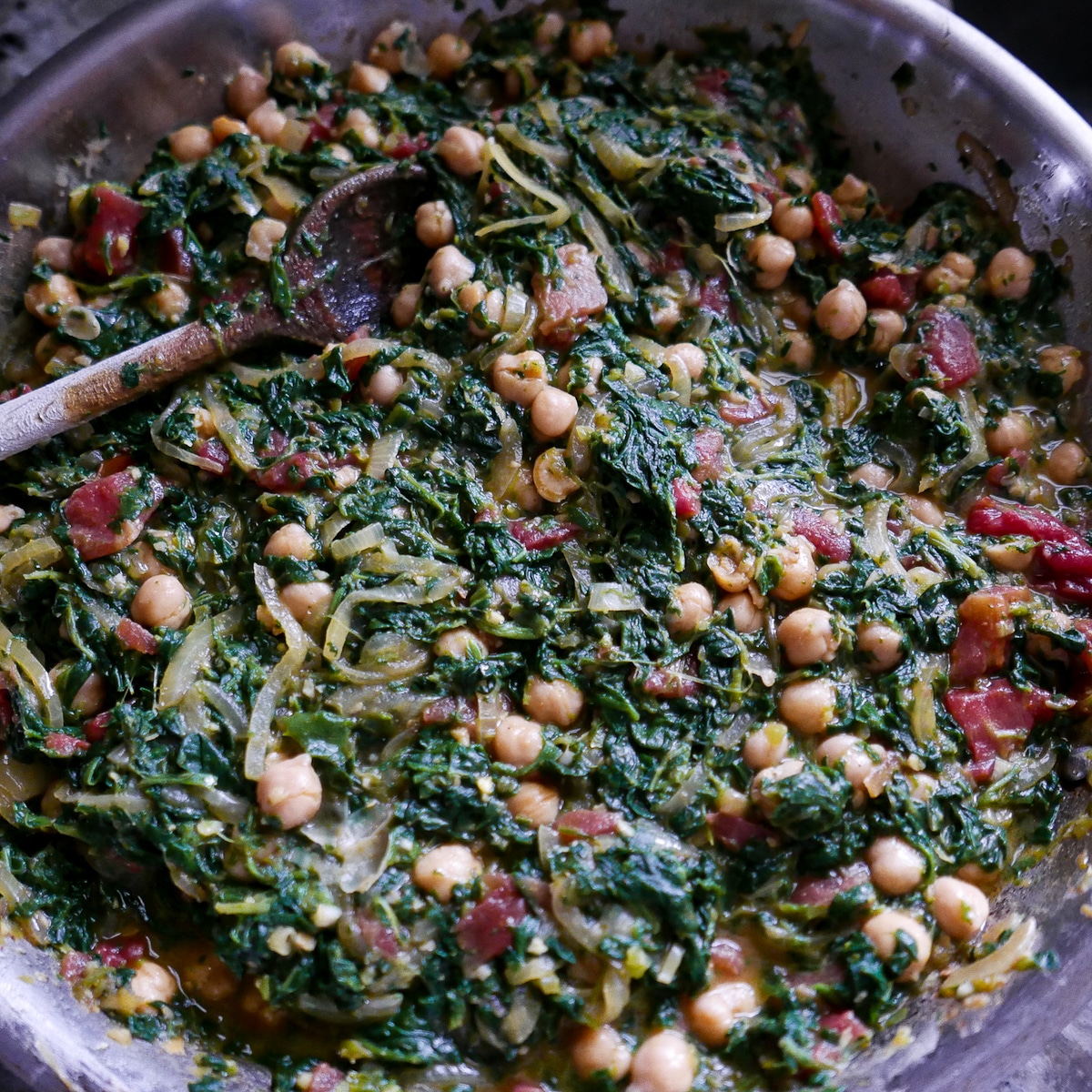 skillet with chickpeas, spinach, tomatoes, and onion and a wooden spoon.