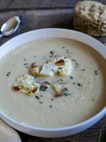 bowl of creamy roasted cauliflower soup on a wooden table with biscuit and spoons in background