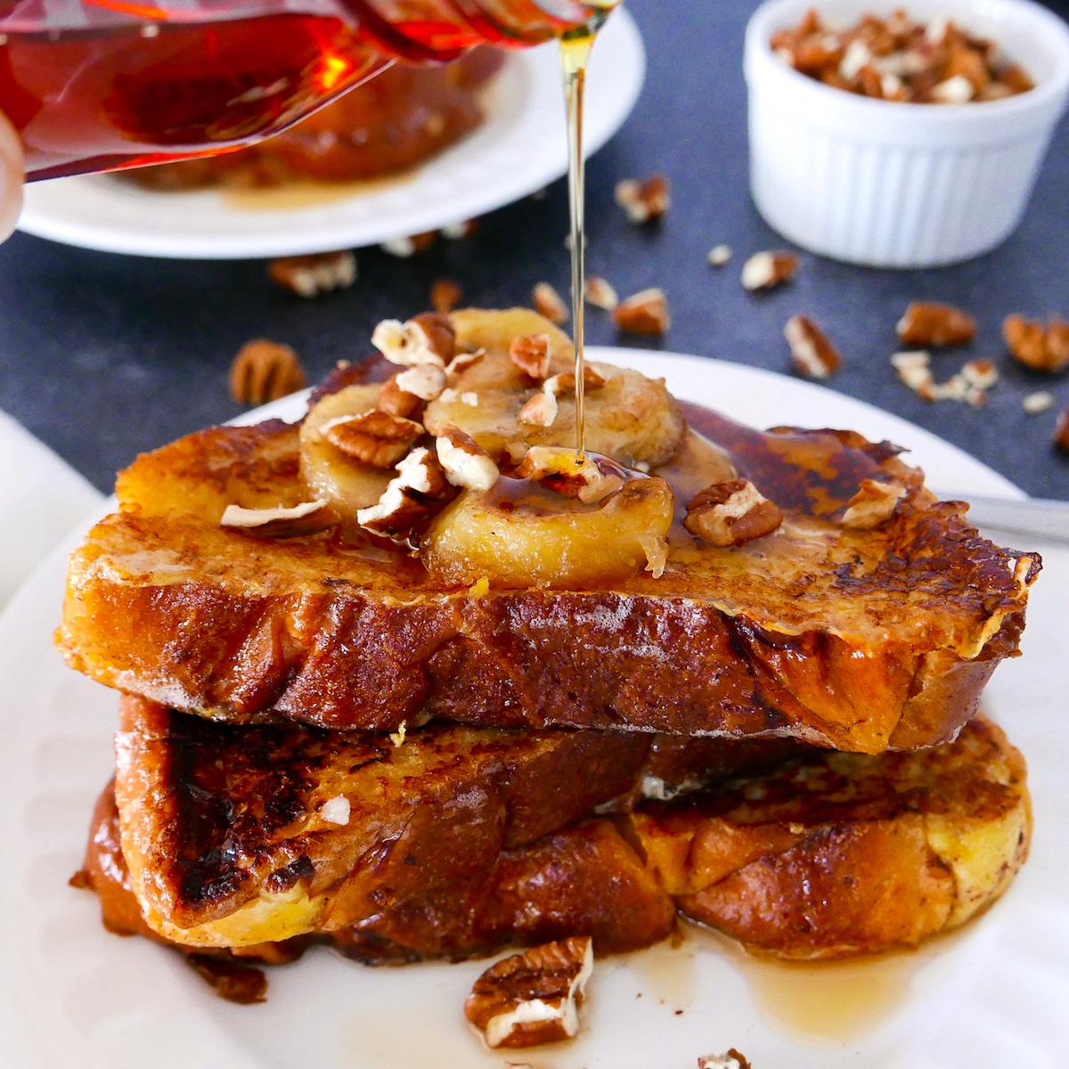 french-toast-with-caramelized-bananas-and-maple-syrup.jpg french toast with caramelized bananas on a white plate with syrup being poured on top.