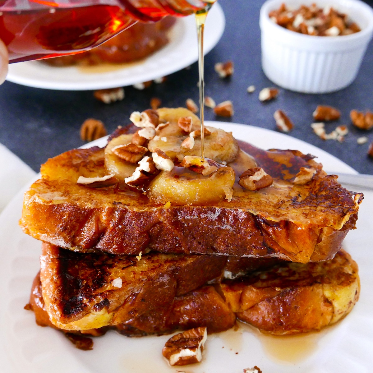 french toast with caramelized bananas on a white plate with syrup being poured on top.