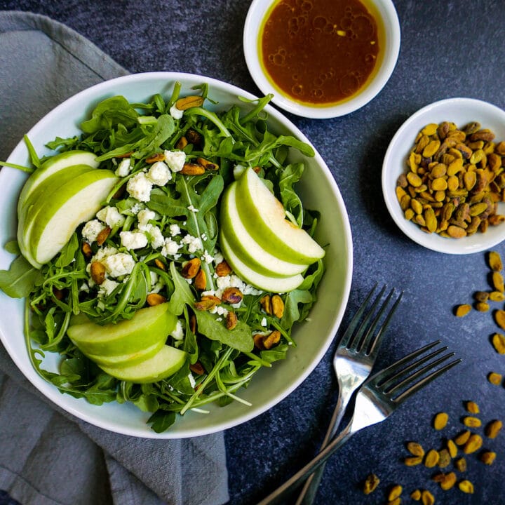 harvest apple salad in a white bowl with cups of vinaigrette and pistachios on table.