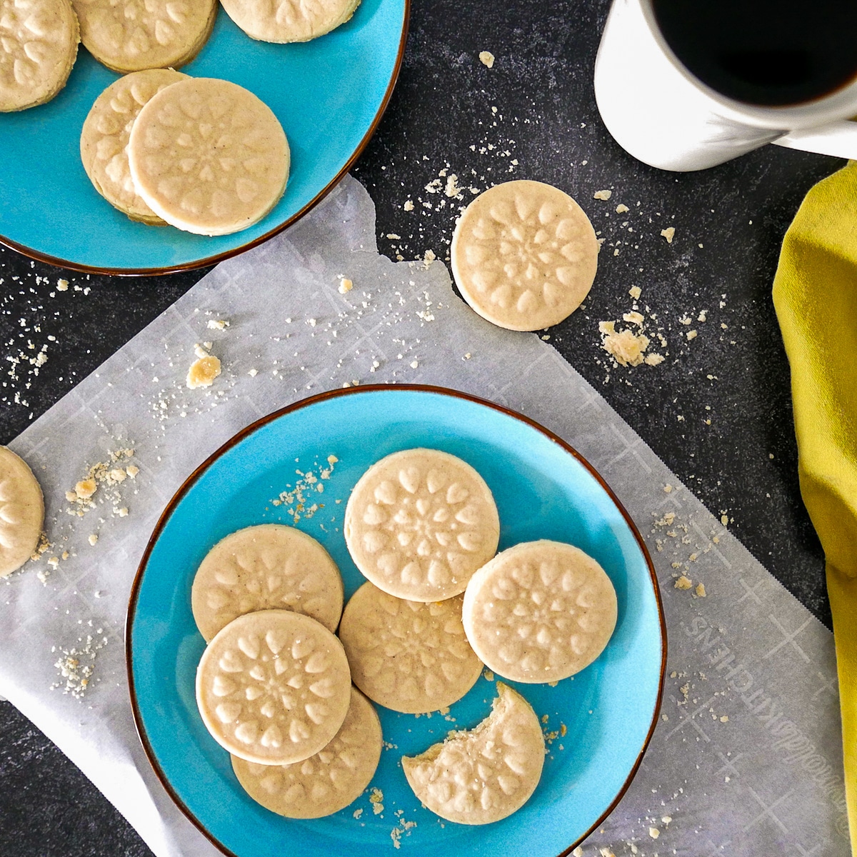 cardamom shortbread cookies arranged on two blue plates with a cup of coffee and cookie crumbs on table.