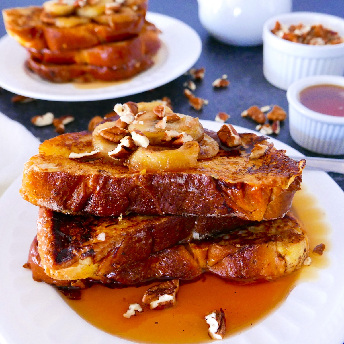 Two plates of banana french toast topped with toasted pecans and maple syrup.