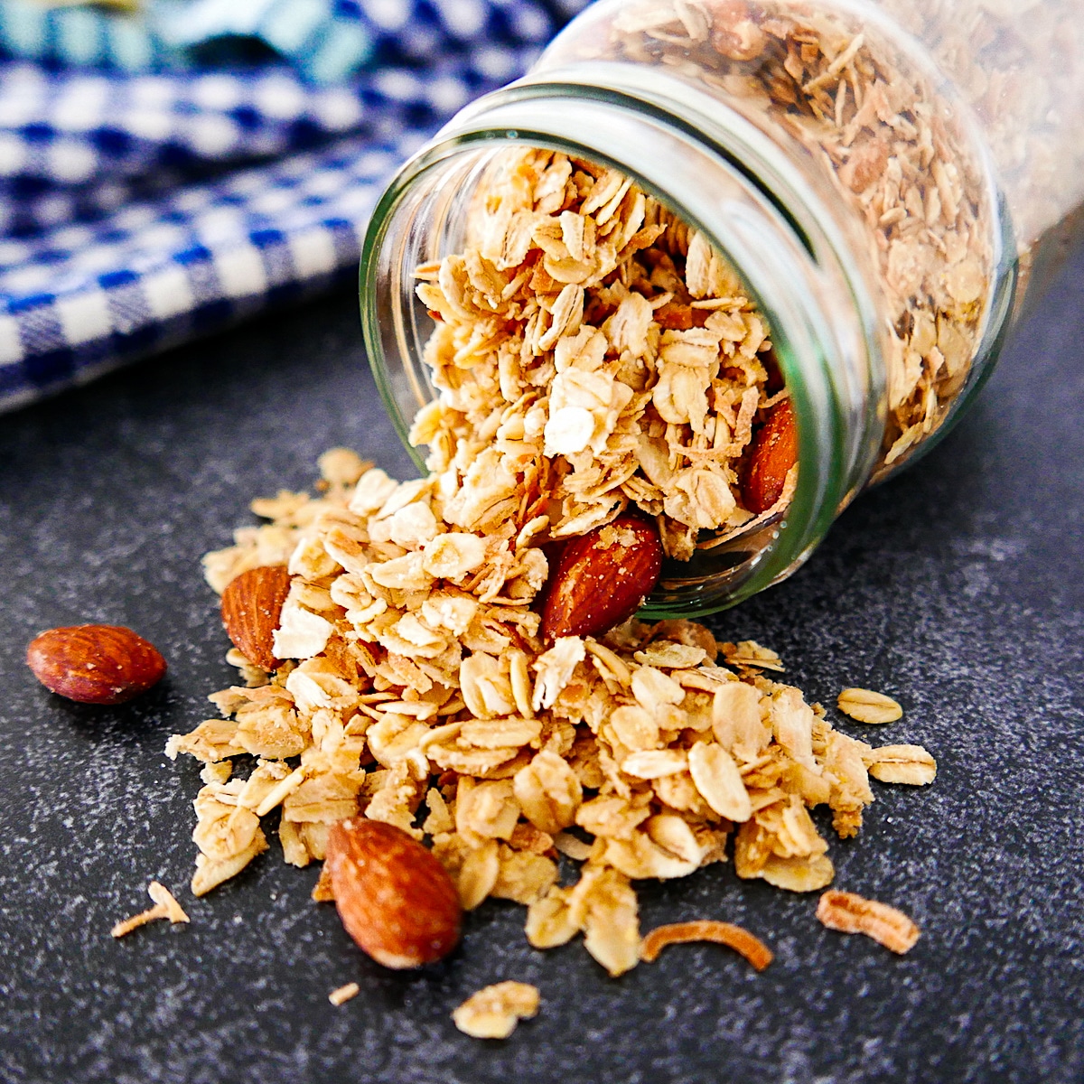 Granola spilling out of a glass jar with a gingham napkin in the background.